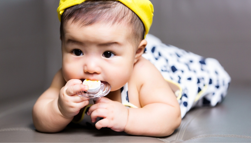 Cute happy asian baby sucking nipple pacifier laying on sofa, 5 months after birth